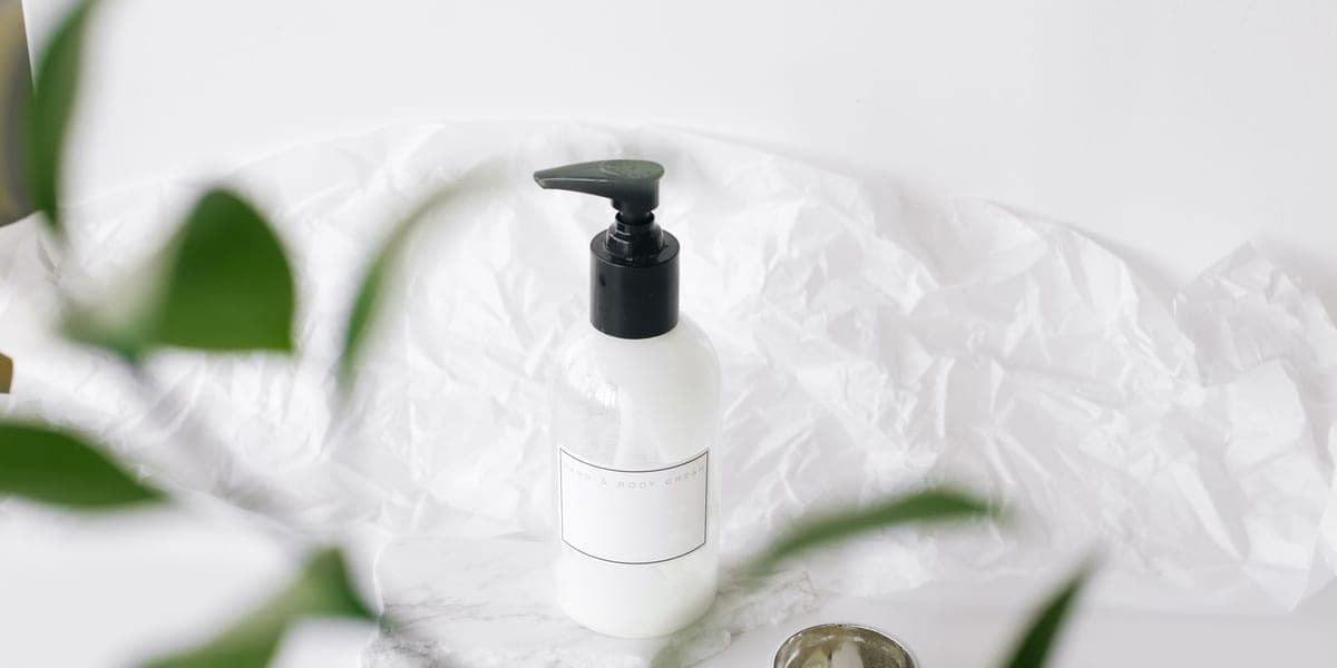 A white pump bottle on a pale surface framed by green leaves.