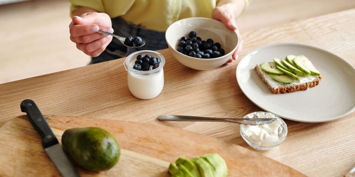 A person adding blueberries to yogurt beside avocado toast on a wooden table.
