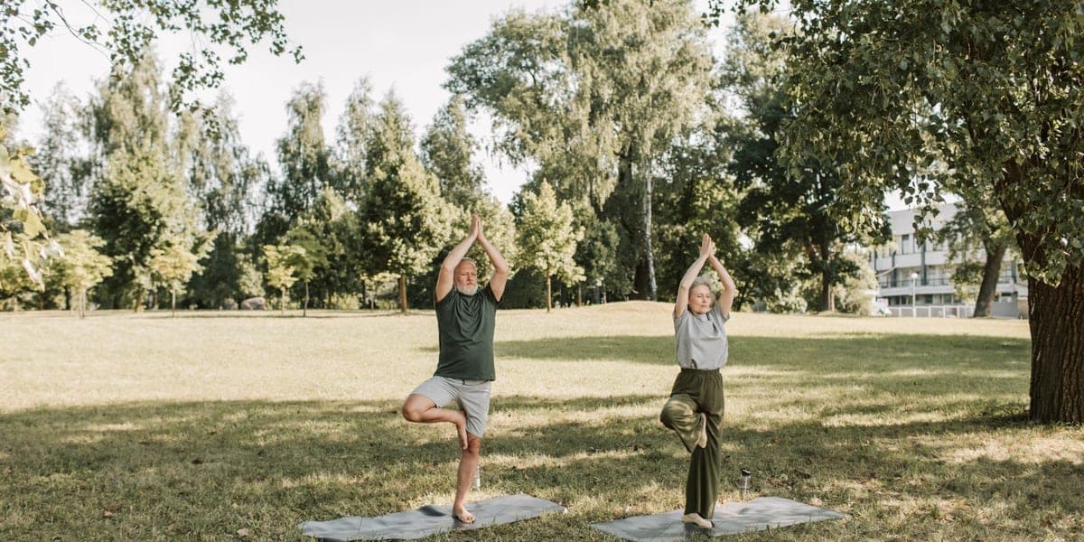 Two older adults practicing standing yoga poses on mats in a park.