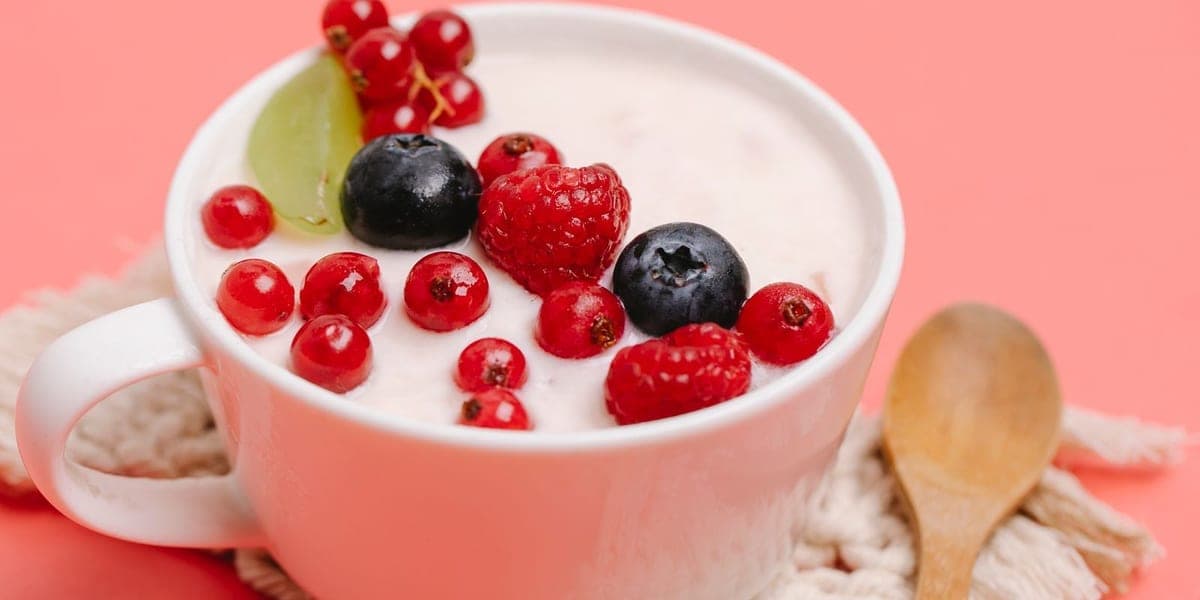 White mug of yogurt topped with berries beside a wooden spoon on a pink surface
