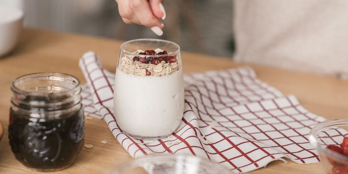 A hand adding toppings to a glass of yogurt with oats and pomegranate seeds