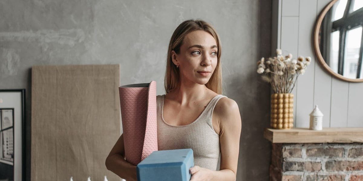 A woman holds a rolled pink yoga mat and two blue foam blocks indoors
