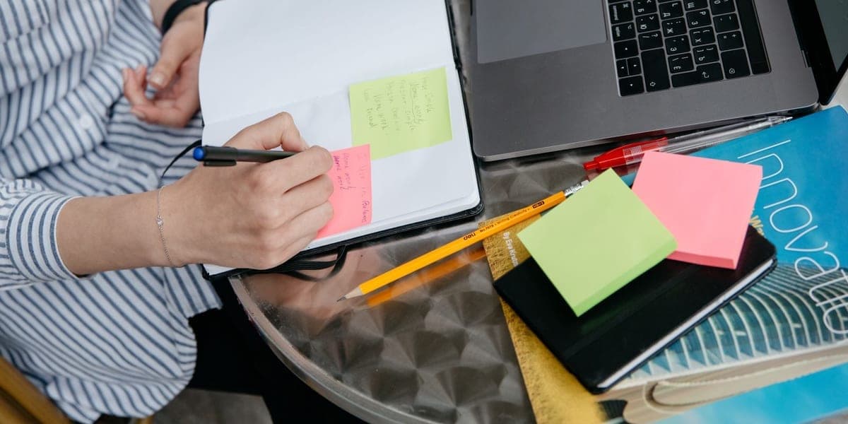 A person writing notes in an open notebook beside a laptop