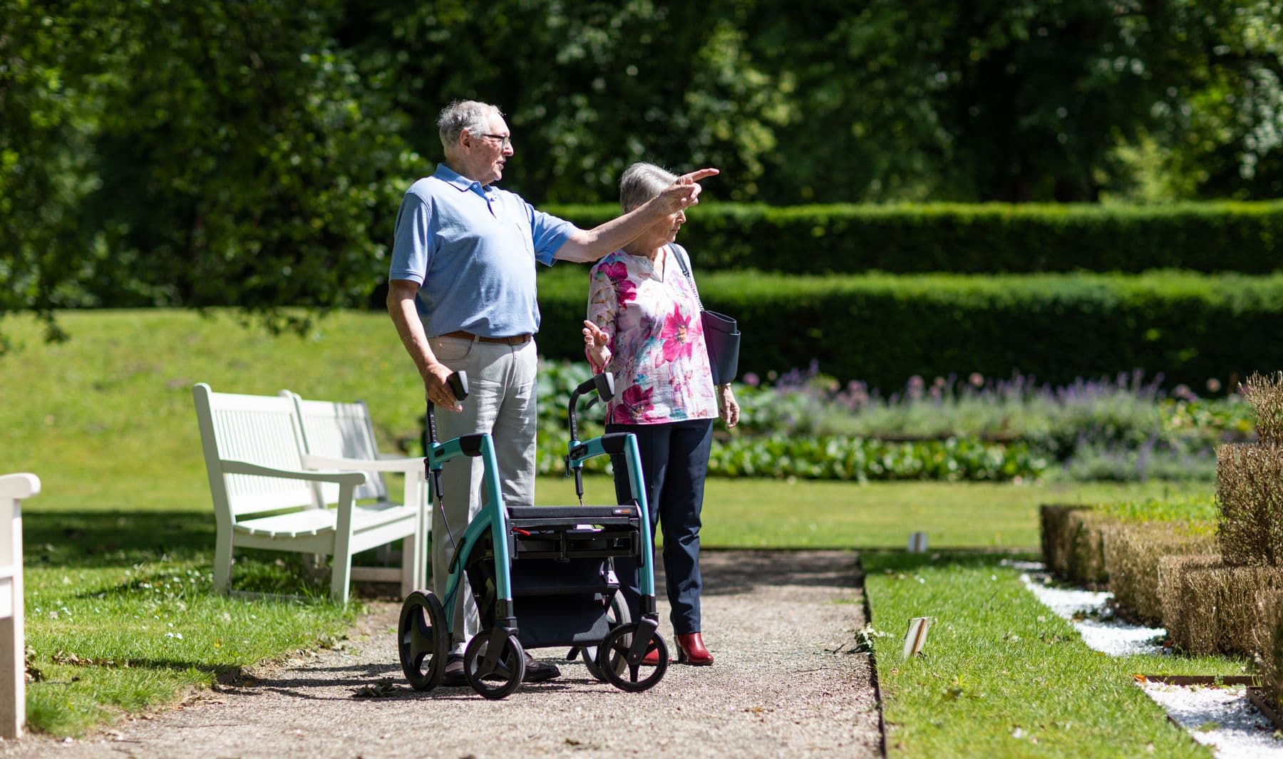 Two older adults with a wheeled walker enjoying a sunny garden path.