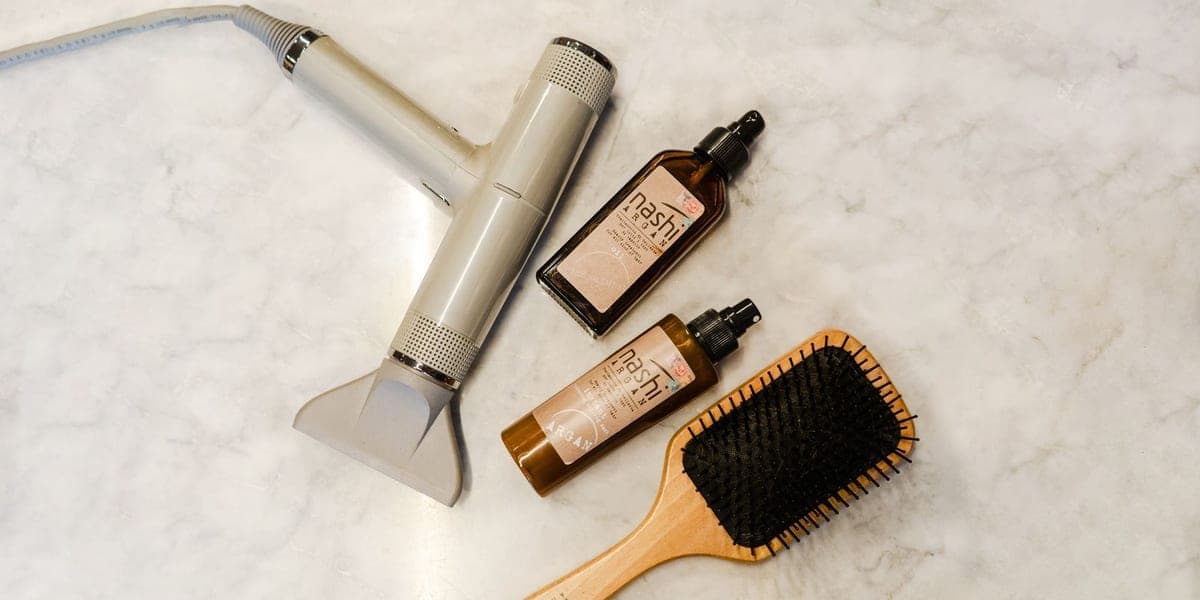 A hair dryer, wooden brush and two hair-care bottles on a pale marble surface