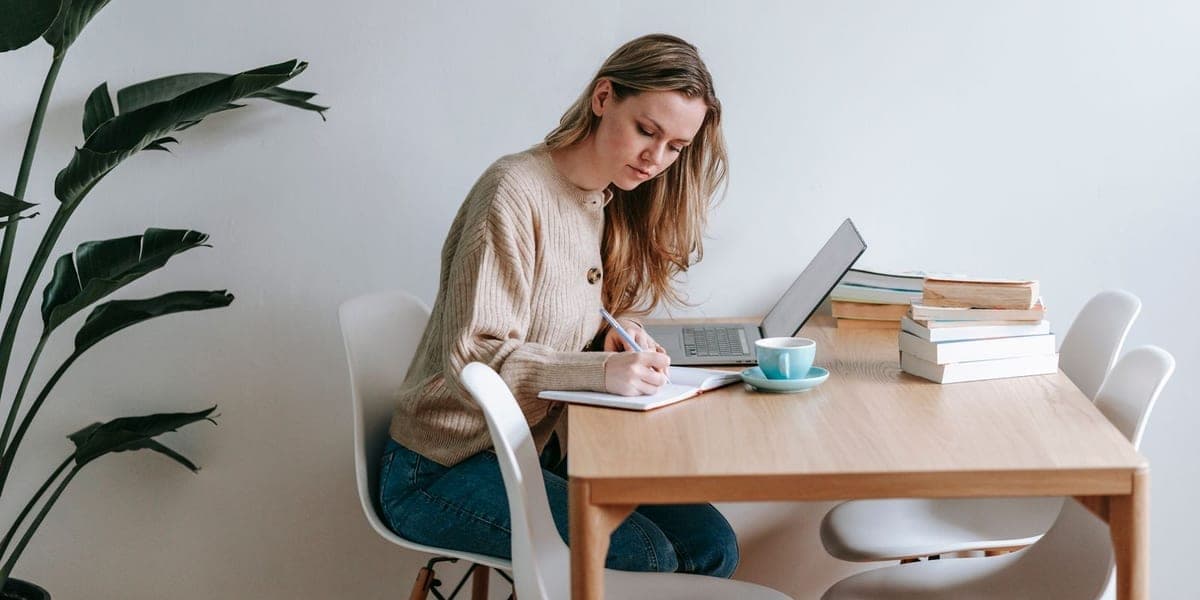 A woman writing in a notebook at a table beside a laptop.