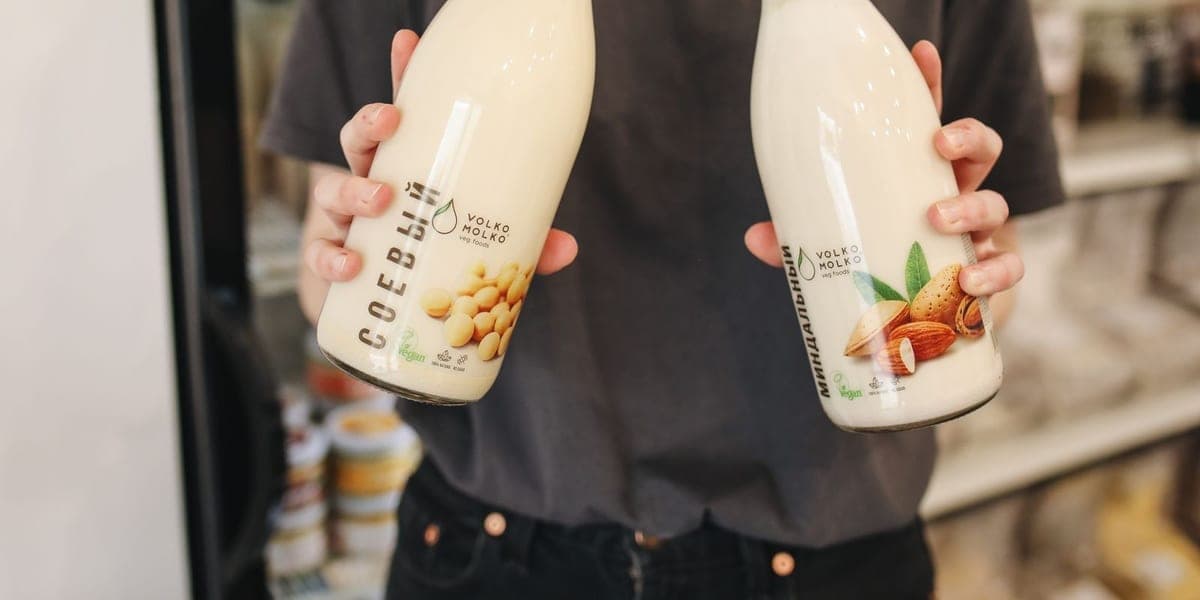 Hands holding two glass bottles of plant milk in a grocery store.