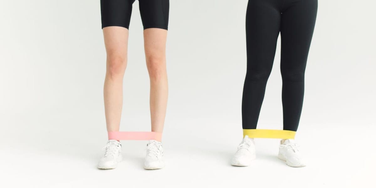 Two people standing with pink and yellow loop resistance bands around their lower legs