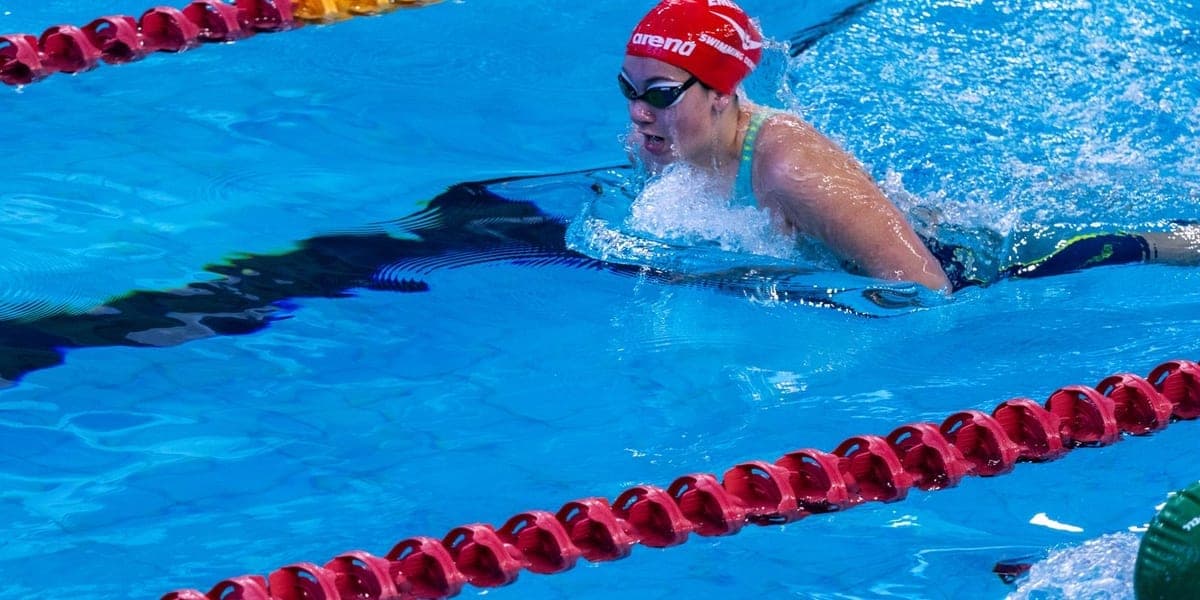 A swimmer wearing goggles and a red cap moving between lane ropes in a pool.