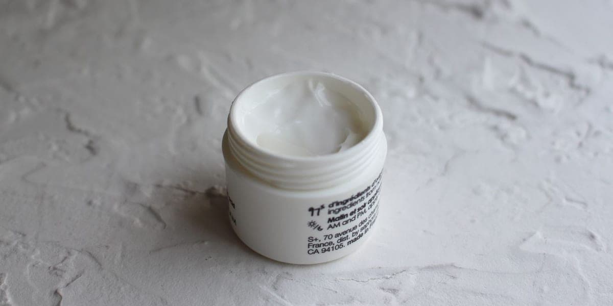 An open white jar of face cream on a pale textured surface