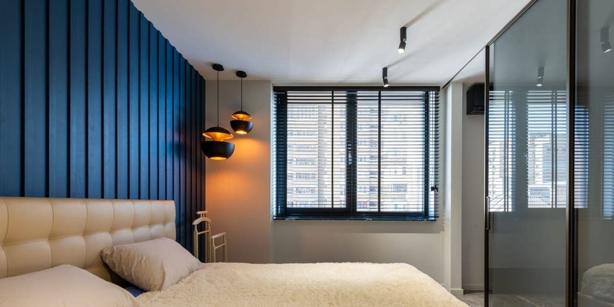 A bed with pale pillows and a cream cover beside a blue wall and window blinds.