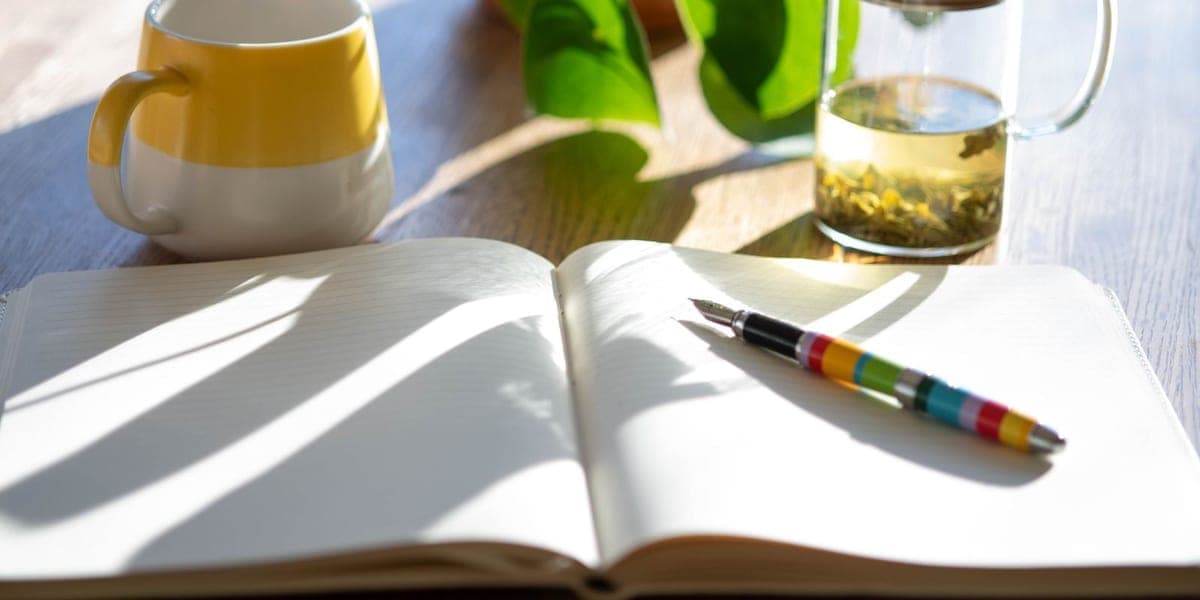 An open notebook and colorful pen beside a mug and glass teapot in morning sunlight.