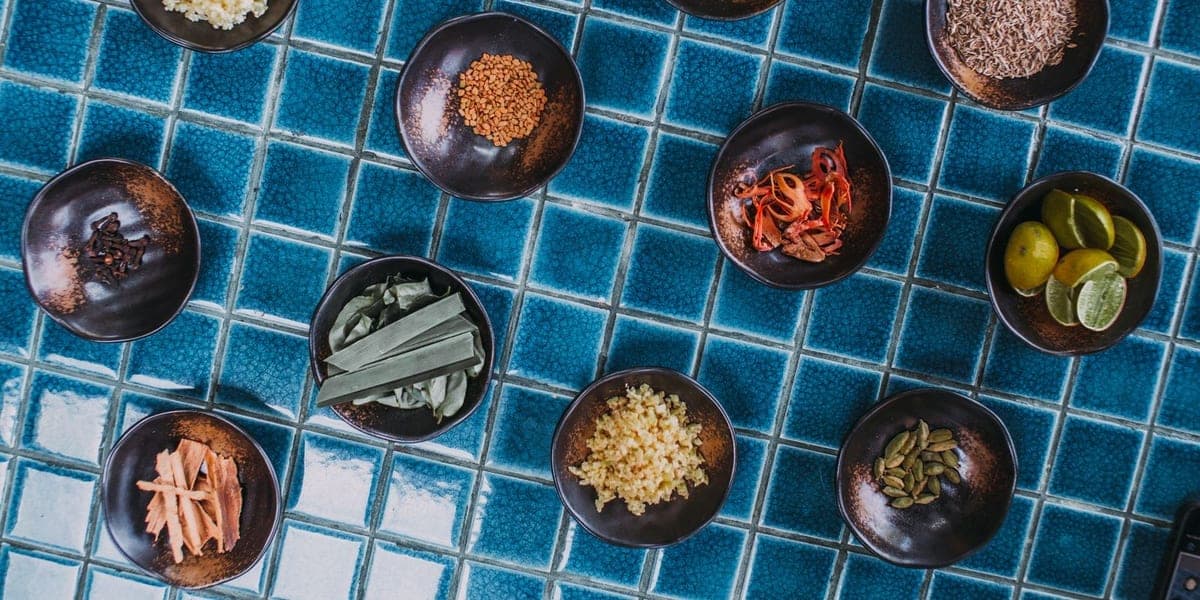 Bowls of spices, aromatic ingredients and lime arranged on blue tiles.